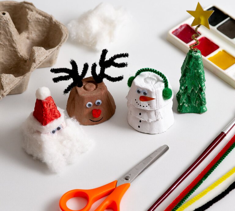 Vier weihnachtliche Figuren aus Eierkarton – Nikolaus, Rentier, Schneemann und Tannenbaum – stehen auf hellem Basteltisch. | © Shutterstock/ Diana Lit