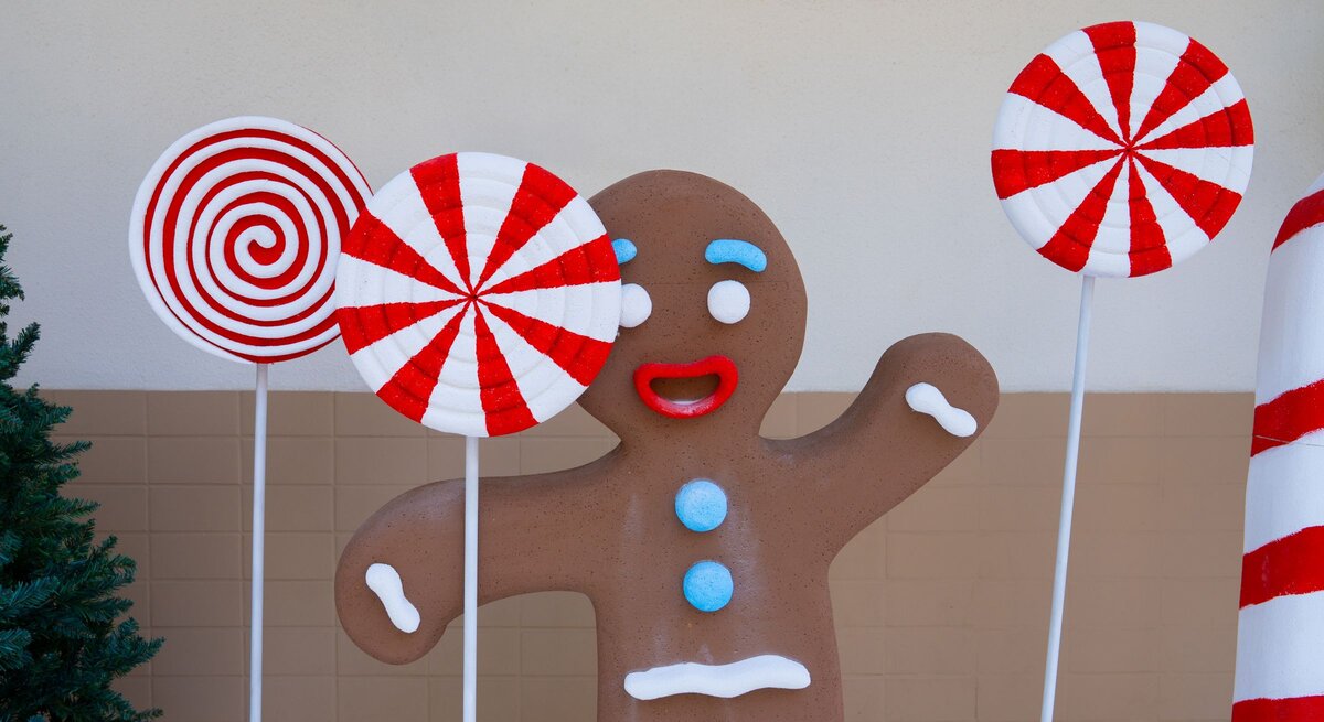 Fröhliche Lebkuchenfigur mit roten Lippen und blauen Knöpfen steht zwischen riesigen rot-weißen Zuckerstangen-Lollis.