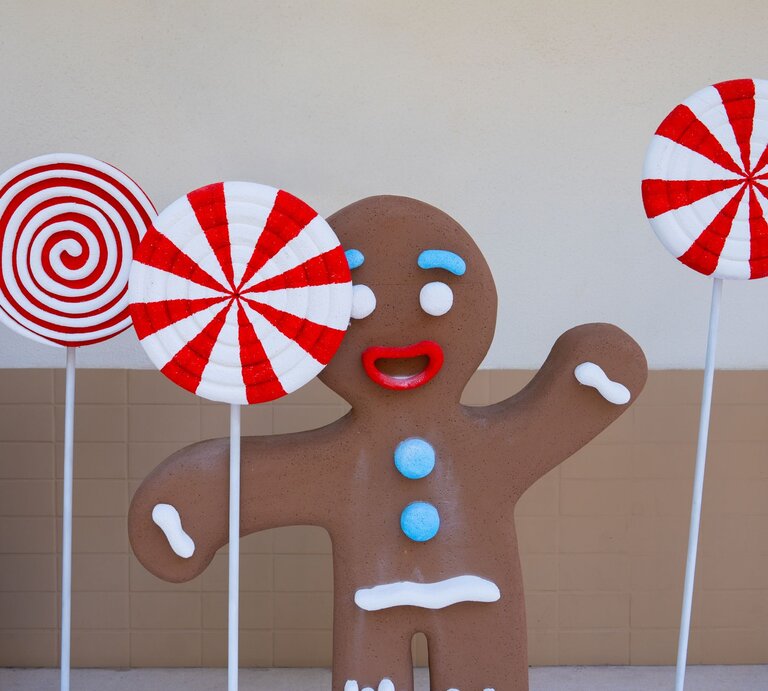 Fröhliche Lebkuchenfigur mit roten Lippen und blauen Knöpfen steht zwischen riesigen rot-weißen Zuckerstangen-Lollis. | © Shutterstock/Sompaen