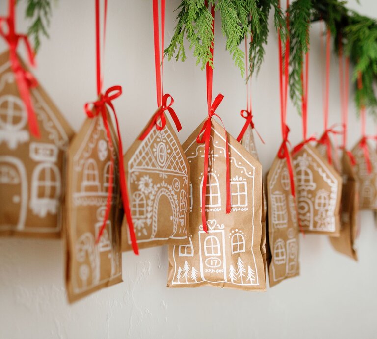 Zwanzig kleine Lebkuchenhäuschen mit weißem Muster hängen als Girlande an roten Bändern unter Tannenzweigen. | © Shutterstock/ Erin Egleston Photography/Canva/Collage:bildderfrau.de