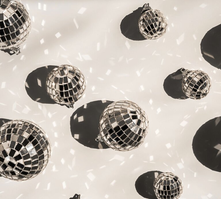 Mehrere glänzende Discokugeln werfen tanzende Lichtreflexe und runde Schatten auf einen hellen Untergrund. | © Shutterstock/Zhenny-zhenny