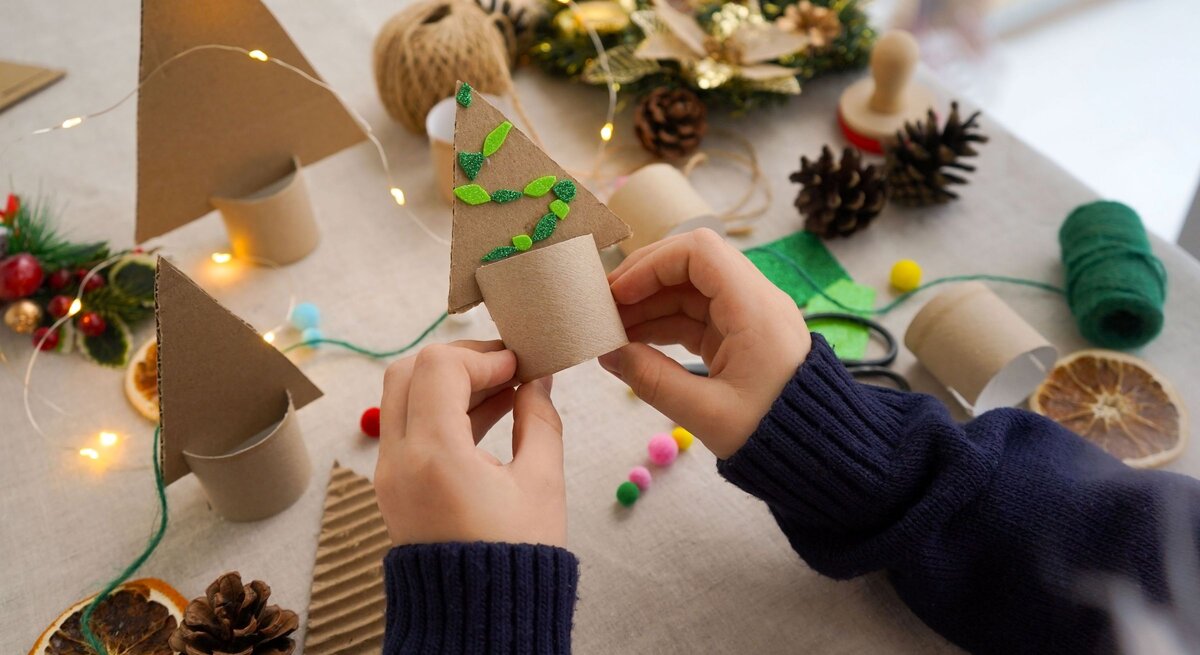 Weihnachtliche Teelichthalter mit Zapfen, Filz, Lichterkette und Trockenobst, Kind hält fertigen Baum aus Klopapierrollen in den Händen.