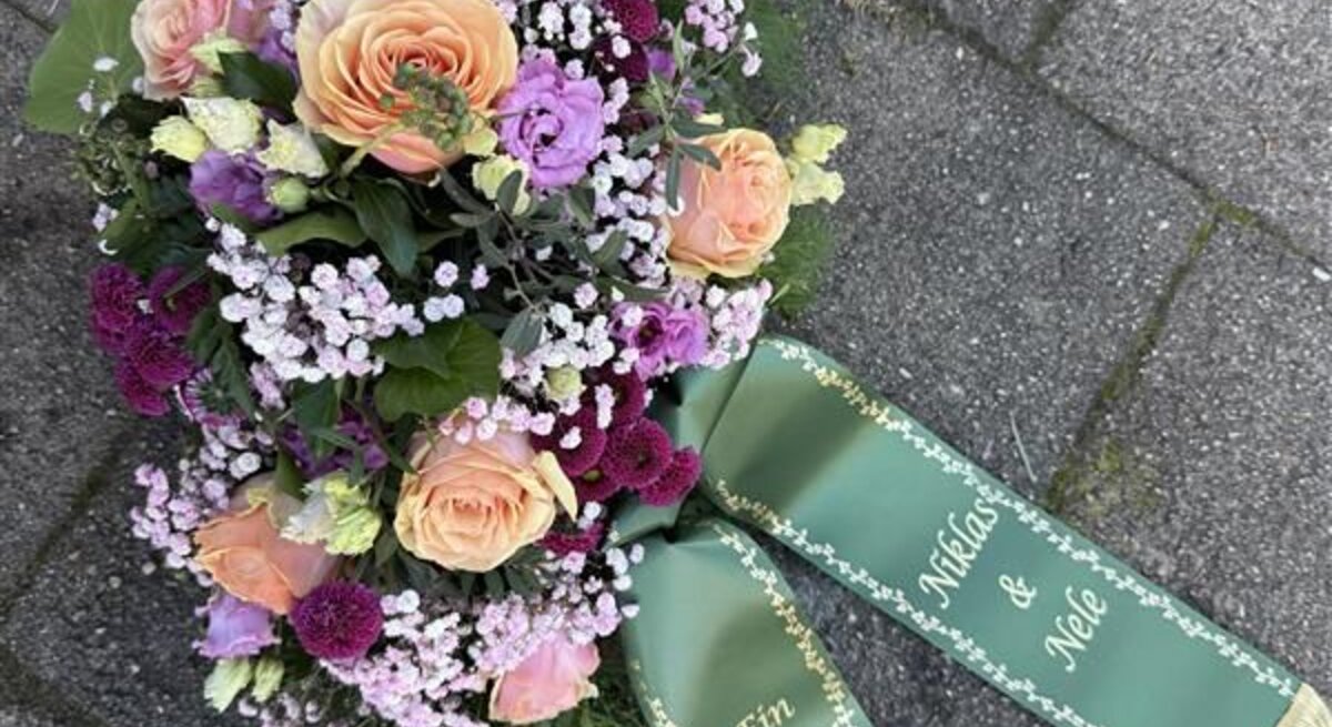 Ein buntes Grabgesteck mit Rosen, Nelken und Schleierkraut liegt mit grünem Schleifenband auf grauen Pflastersteinen.