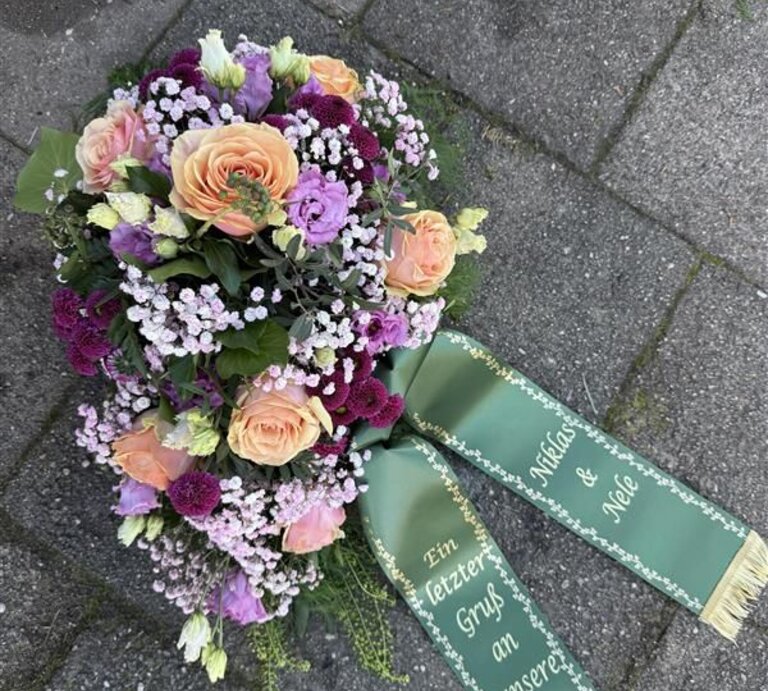 Ein buntes Grabgesteck mit Rosen, Nelken und Schleierkraut liegt mit grünem Schleifenband auf grauen Pflastersteinen. | © Madeleine Liedke 