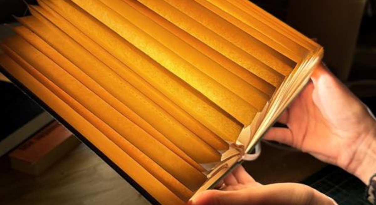 Mehrere kunstvoll gefaltete, warm leuchtende Buch-Lampen aus alten Seiten liegen auf einem Holztisch, eine wird gerade in Händen gehalten.