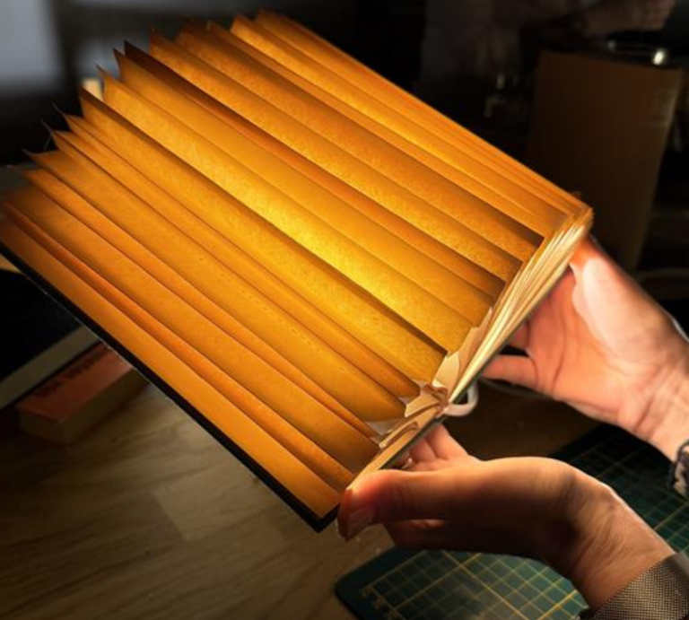 Mehrere kunstvoll gefaltete, warm leuchtende Buch-Lampen aus alten Seiten liegen auf einem Holztisch, eine wird gerade in Händen gehalten. | © Canva/Collage:bildderfrau.de