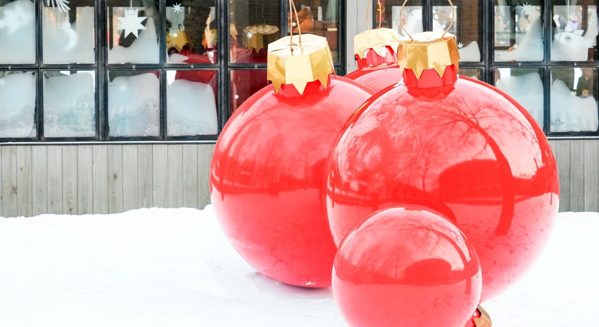 Vier übergroße rote Christbaumkugeln mit goldenen Aufhängern stehen dekorativ im Schnee vor einem Fenster mit Eisblumen.