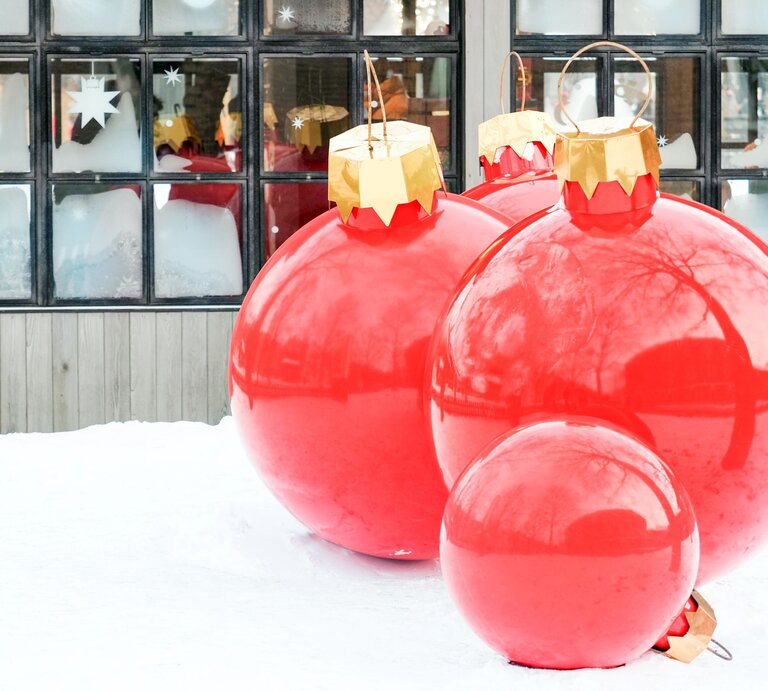 Vier übergroße rote Christbaumkugeln mit goldenen Aufhängern stehen dekorativ im Schnee vor einem Fenster mit Eisblumen. | © Shutterstock/ANGHI