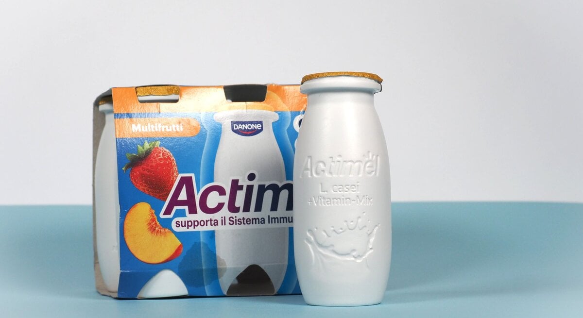 Eine weiße Actimel-Flasche mit goldener Versiegelung steht vor einem bunten Viererpack in Multifrucht-Design auf blauem Untergrund.
