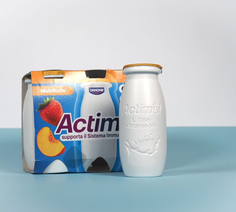 Eine weiße Actimel-Flasche mit goldener Versiegelung steht vor einem bunten Viererpack in Multifrucht-Design auf blauem Untergrund. | © Claudio Divizia - stock.adobe.com