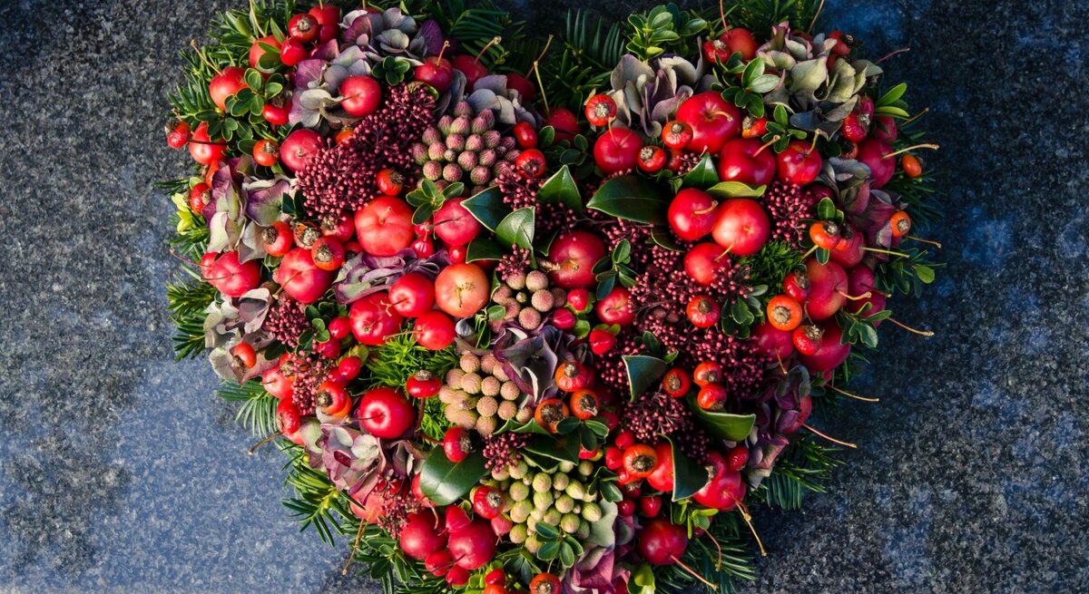Herzförmiges Grabgesteck für Totensonntag  aus roten Beeren, Hortensien, Blättern und Tannenzweigen auf dunkelgrauem Naturstein.