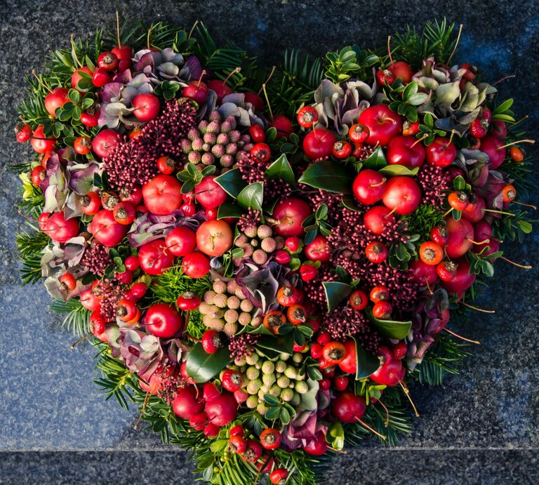 Herzförmiges Grabgesteck für Totensonntag  aus roten Beeren, Hortensien, Blättern und Tannenzweigen auf dunkelgrauem Naturstein. | © Shutterstock/ katarinag