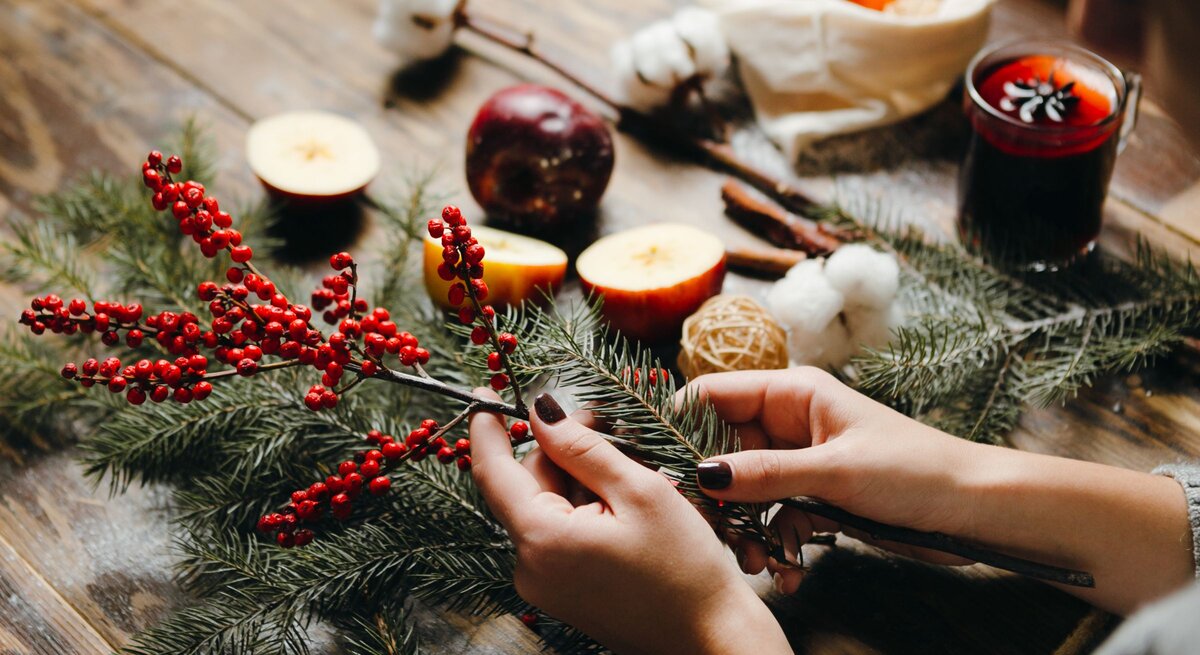 Zwei Hände mit dunkel lackierten Nägeln arrangieren Tannenzweige, Thuja und rote Beeren auf einem rustikalen Holztisch.