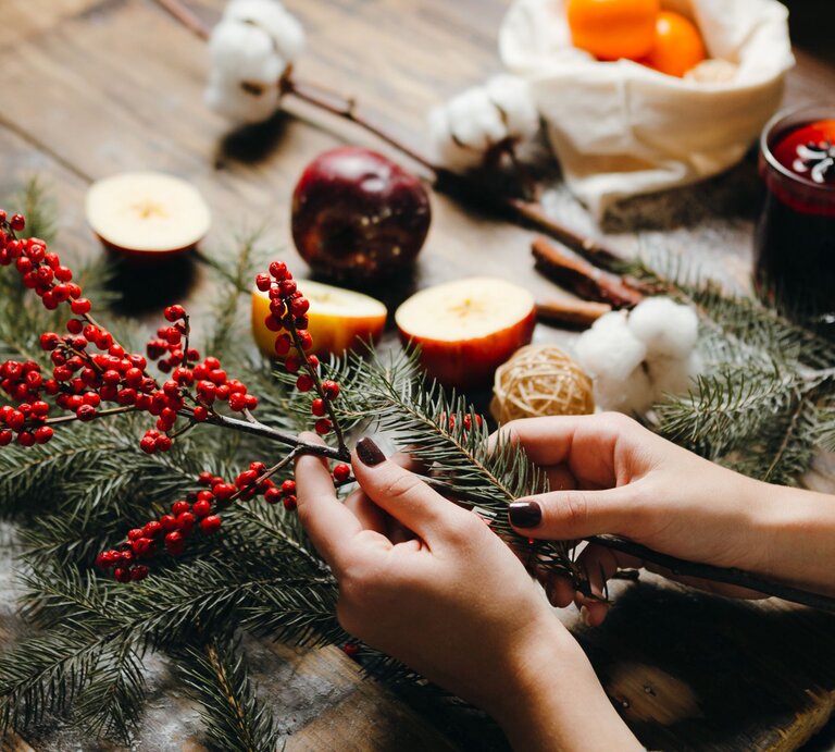 Zwei Hände mit dunkel lackierten Nägeln arrangieren Tannenzweige, Thuja und rote Beeren auf einem rustikalen Holztisch. | © Shutterstock/Chester-Alive