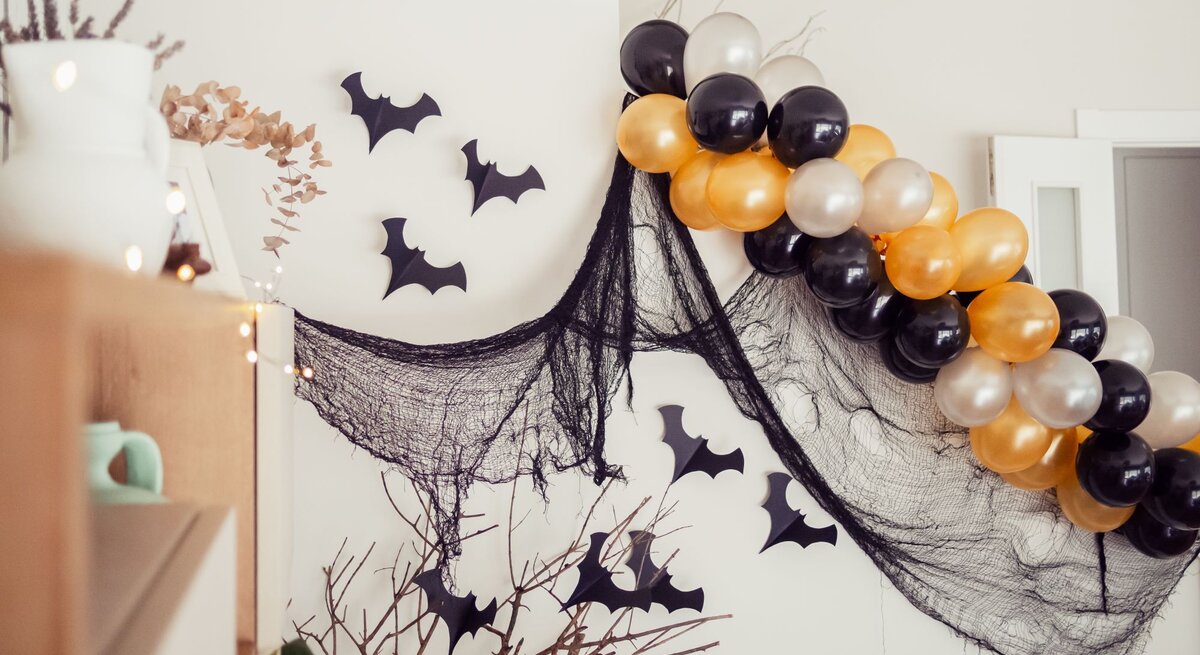 Halloween-Deko mit schwarzen Stoffnetzen, Papierfledermäusen und glänzenden Luftballons in Schwarz, Gold und Perlweiß.