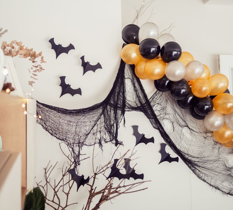 Halloween-Deko mit schwarzen Stoffnetzen, Papierfledermäusen und glänzenden Luftballons in Schwarz, Gold und Perlweiß. | © Shutterstock/Valentina Musumeci