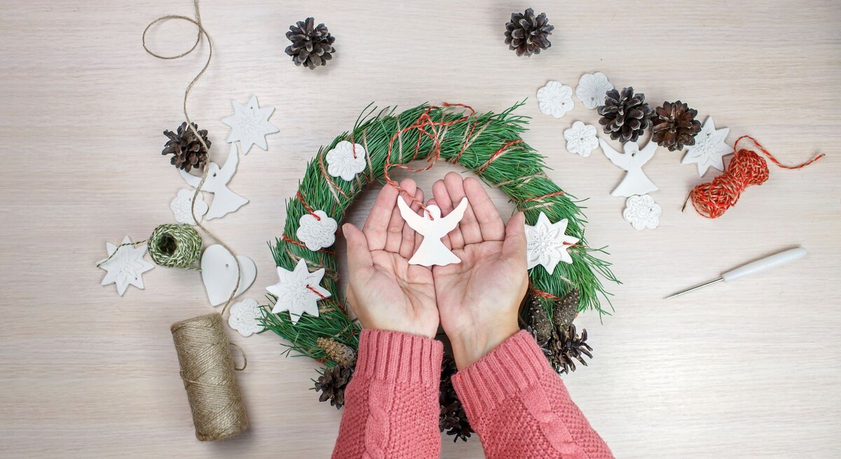 Zwei Hände in rosa Strick halten einen weißen Engel aus Salzteig über einem festlich geschmückten Adventskranz.