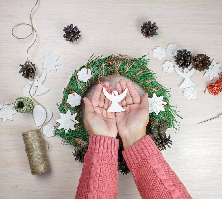 Zwei Hände in rosa Strick halten einen weißen Engel aus Salzteig über einem festlich geschmückten Adventskranz. | © Shutterstock/Maria Symchych