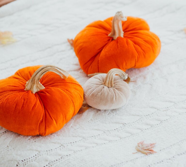 Zwei orangefarbene und ein beiger Zierkürbis aus Strumpfhosen gebastelt, liegen dekorativ auf einer weißen Strickdecke mit Herbstblättern. | © Shutterstock/ reddish
