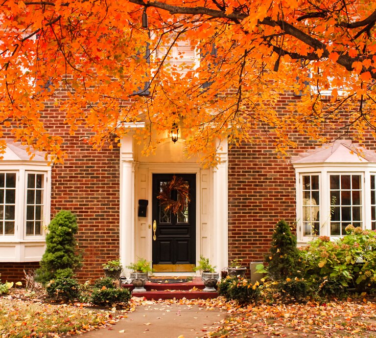 Eingang eines Backsteinhauses mit schwarzer Tür, Herbstkranz und beidseitigen Fenstern, umrahmt von leuchtend orangenen Blättern. | © Adobe Stock/ Susan Vineyard