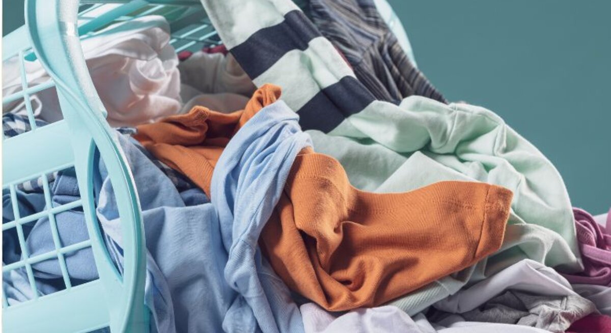 Nahaufnahme eines Korbs mit frisch gefalteten, weichen Handtüchern in Grau und Rosa, gehalten von einer Person in lässiger Kleidung vor neutralem Hintergrund.