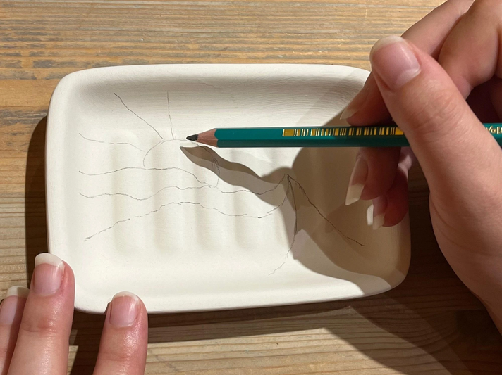 Eine Hand zeichnet mit einem grünen Stift eine Skizze aus Sonne und Bergen in eine unbemalte Keramikschale auf einem Holztisch. | © Kaja Wundersitz/ Linda Pötzsch/ BIld der Frau