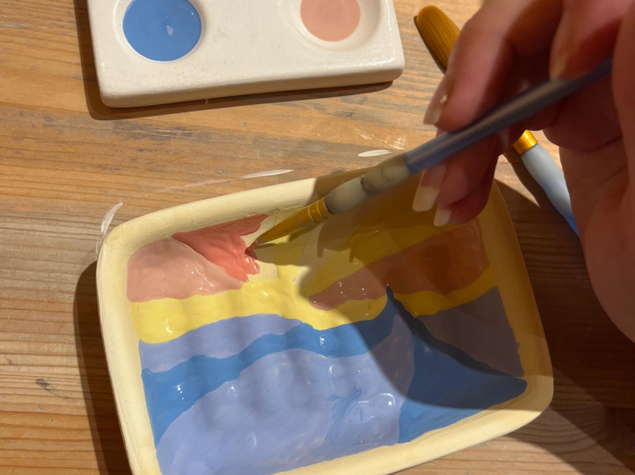 Ein Pinsel malt rosa und gelbe Farben in eine halb bemalte Keramikschale mit einem Landschaftsmotiv auf einem Holztisch. | © Kaja Wundersitz/ Linda Pötzsch/ BIld der Frau