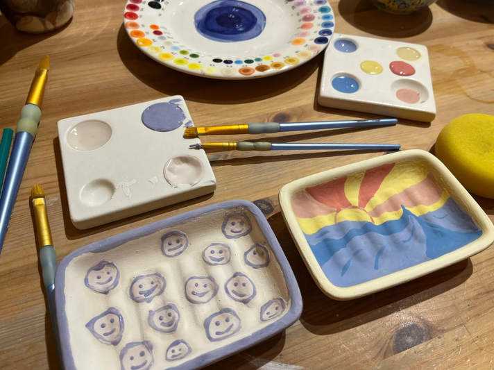 Zwei bemalte Keramikschalen mit Smileys und Sonnenmotiv liegen neben Farbtellern, Pinseln und einem gelben Schwamm auf Holztisch. | © Kaja Wundersitz/ Linda Pötzsch/ BIld der Frau