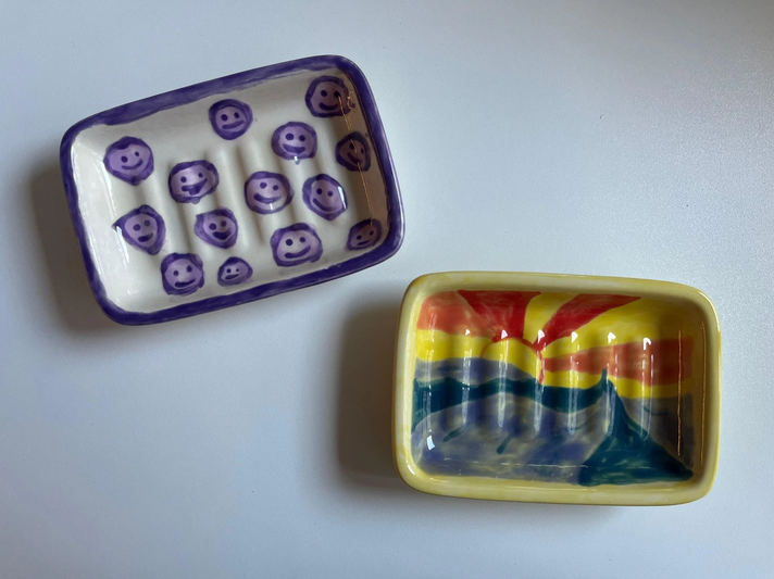 Zwei bemalte Keramikschalen, eine mit lila Smileys und die andere mit einem farbenfrohen Sonnenaufgang-Motiv, liegen auf weißer Oberfläche. | © Kaja Wundersitz/ Linda Pötzsch/ BIld der Frau
