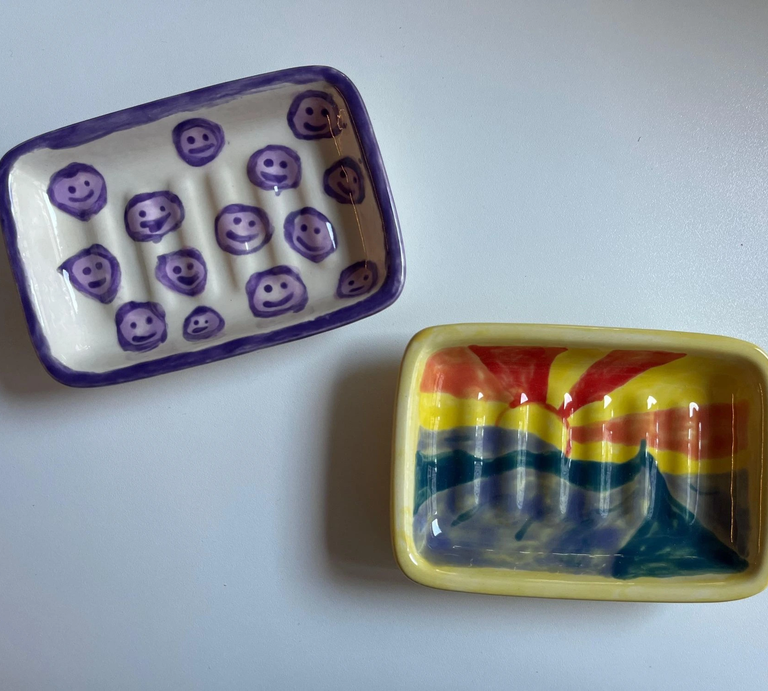 Zwei bemalte Keramikschalen, eine mit lila Smileys und die andere mit einem farbenfrohen Sonnenaufgang-Motiv, liegen auf weißer Oberfläche. | © Kaja Wundersitz/ Linda Pötzsch/ BIld der Frau