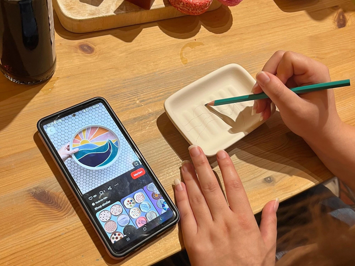 Eine Hand zeichnet mit einem grünen Stift ein Wellenmotiv in eine Keramikschale, daneben ein Smartphone mit ähnlicher Vorlage. | © Kaja Wundersitz/ Linda Pötzsch/ BIld der Frau