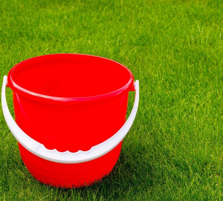 Ein leerer, knallroter Plastikeimer mit weißem Henkel steht auf dichtem, frisch gemähtem, sattem grünen Rasen. | © AdobeStock/ArtCookStudio