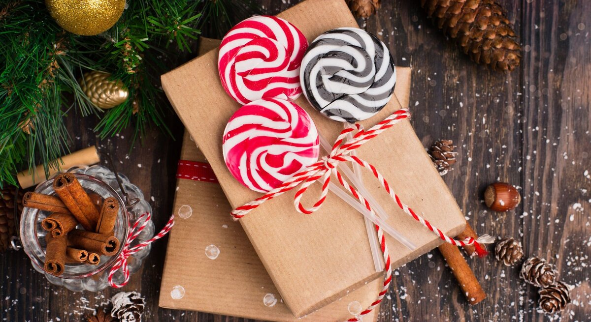 Zwei mit rotem Band dekorierte Gutschein-Geschenke, bunte Lollis und Zimtstangen liegen auf dunklem Holzboden, umgeben von Tannenzapfen.