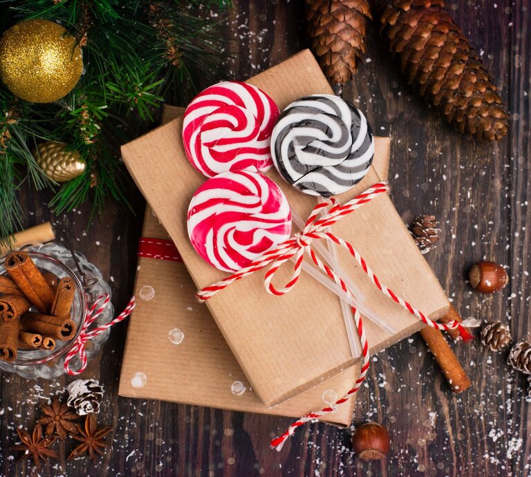 Zwei mit rotem Band dekorierte Gutschein-Geschenke, bunte Lollis und Zimtstangen liegen auf dunklem Holzboden, umgeben von Tannenzapfen. | © Adobe Stock/ Iuliia Metkalova