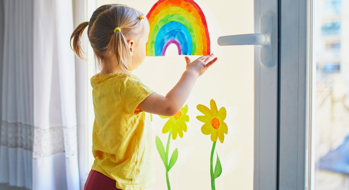 Kleines Mädchen mit blonden Zöpfen betrachtet fröhlich einen bunten Regenbogen am Fenster neben gemalten Sonnenblumen.