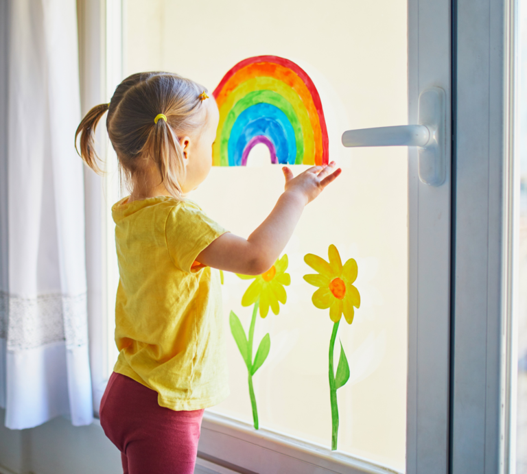 Kleines Mädchen mit blonden Zöpfen betrachtet fröhlich einen bunten Regenbogen am Fenster neben gemalten Sonnenblumen. | © AdobeStock/Ekaterina Pokrovsky