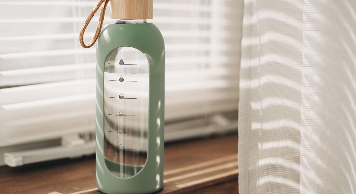 Moderne Glasflasche mit DIY-Wasser-Tracker leuchtet im warmen Licht vor halb geöffneten Jalousien.