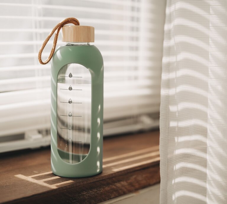 Moderne Glasflasche mit DIY-Wasser-Tracker leuchtet im warmen Licht vor halb geöffneten Jalousien. | © GettyImages/Viktoriya Skorikova