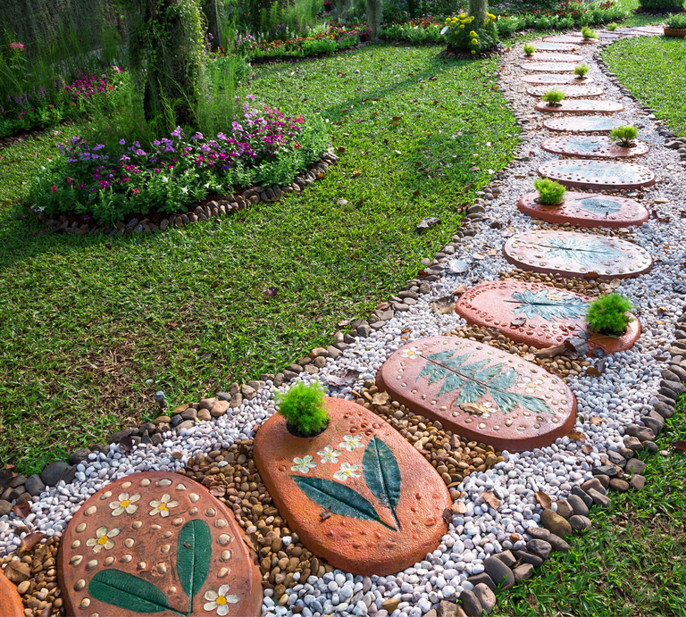 Geschwungener Gartenweg aus bunten Trittsteinen mit Blumenmotiven, umrahmt von Kieseln und gepflegtem Rasen. | © GettyImages/undefined undefined