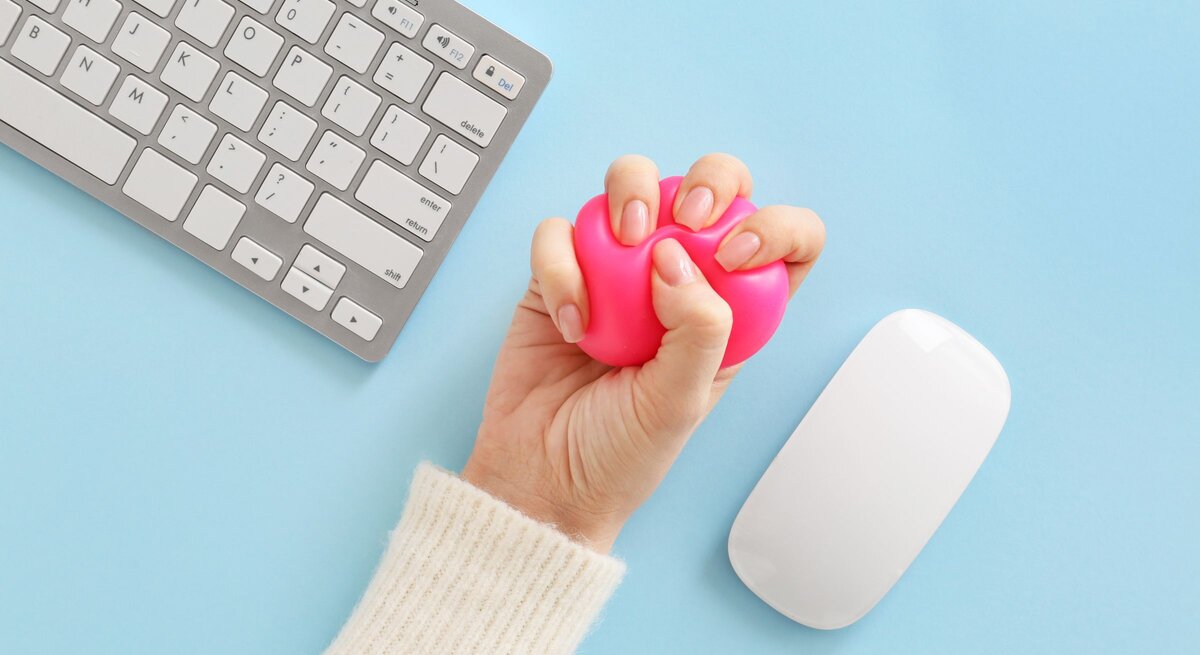 Eine Hand in cremeweißem Strickärmel drückt einen pinken Stressball neben Tastatur und Maus auf hellblauem Untergrund.