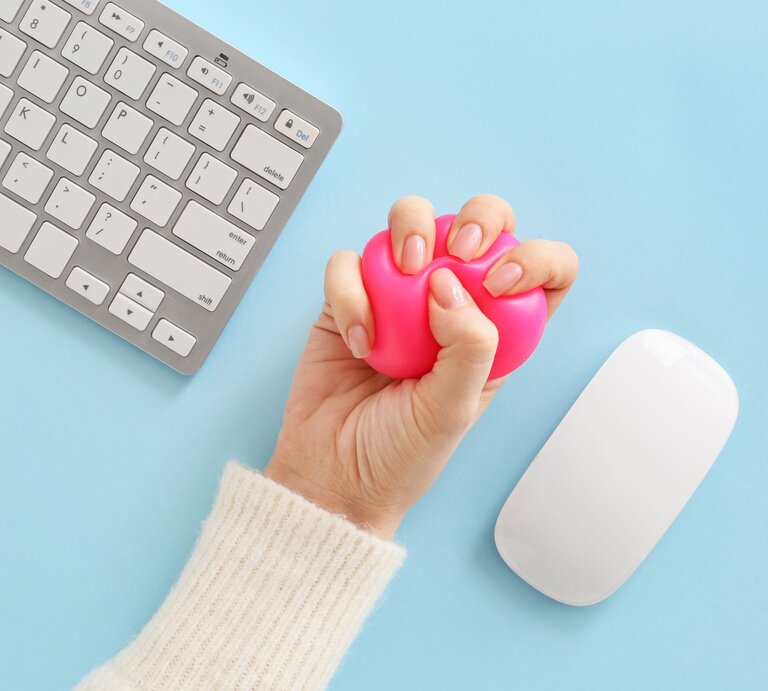 Eine Hand in cremeweißem Strickärmel drückt einen pinken Stressball neben Tastatur und Maus auf hellblauem Untergrund. | © Shutterstock/Pixel-Shot