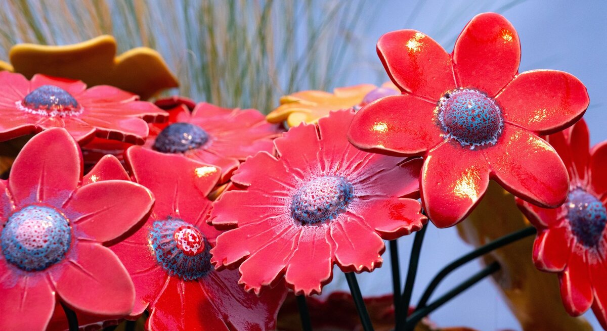 Leuchtend rote DIY-Gartenstecker mit blauen Mitteln glänzen im Sonnenlicht vor unscharfem Gras und blassem Himmel.