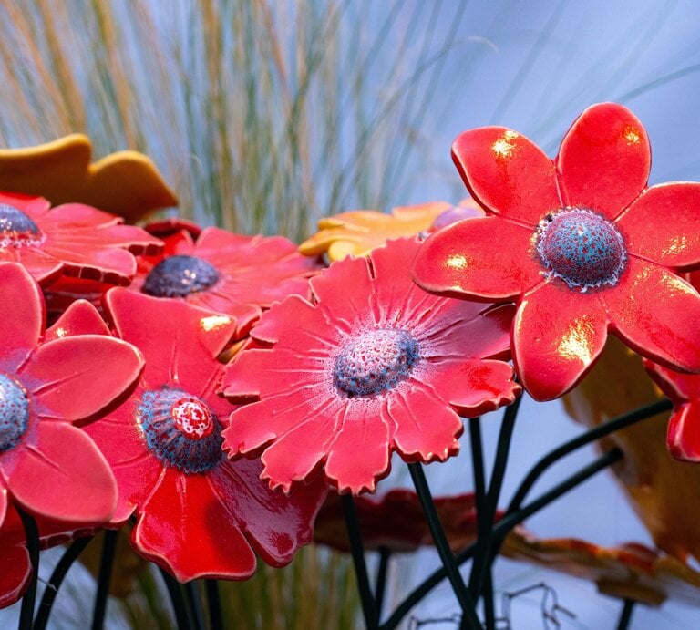 Leuchtend rote DIY-Gartenstecker mit blauen Mitteln glänzen im Sonnenlicht vor unscharfem Gras und blassem Himmel. | © AdobeStock/Comofoto