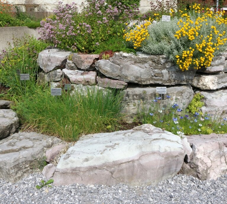 Mehrere Kräuter wachsen in einer Kräuterspirale aus groben Natursteinen vor grauem Schotter und Betonmauer. | © AdobeStock/vulkanismus