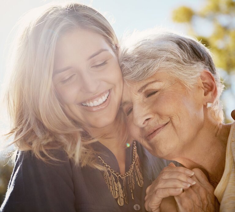 Junge blonde Frau und ältere Frau mit grauem Haar umarmen sich liebevoll am Muttertag und lächeln glücklich im Sonnenlicht eines Gartens. | © AdobeStock/Grady Reese/peopleimages.com