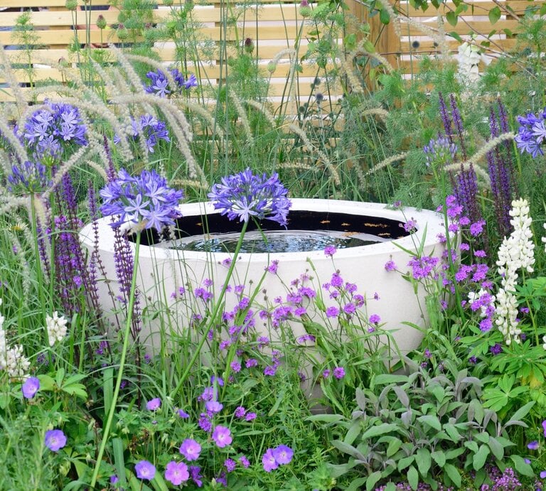 Üppiges Blumenbeet mit violetten und weißen Blüten umgibt ein runder, moderner Solarbrunnen vor hölzerner Sichtschutzwand. | © Adobe Stock/ pauws99