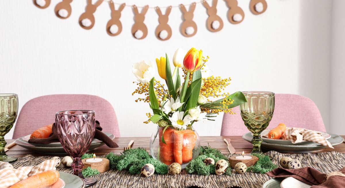 Frühlingshaft gedeckter Tisch mit Tulpenstrauß in Karottenvase, bunten Gläsern, Möhren auf Tellern und Hasengirlande im Hintergrund.