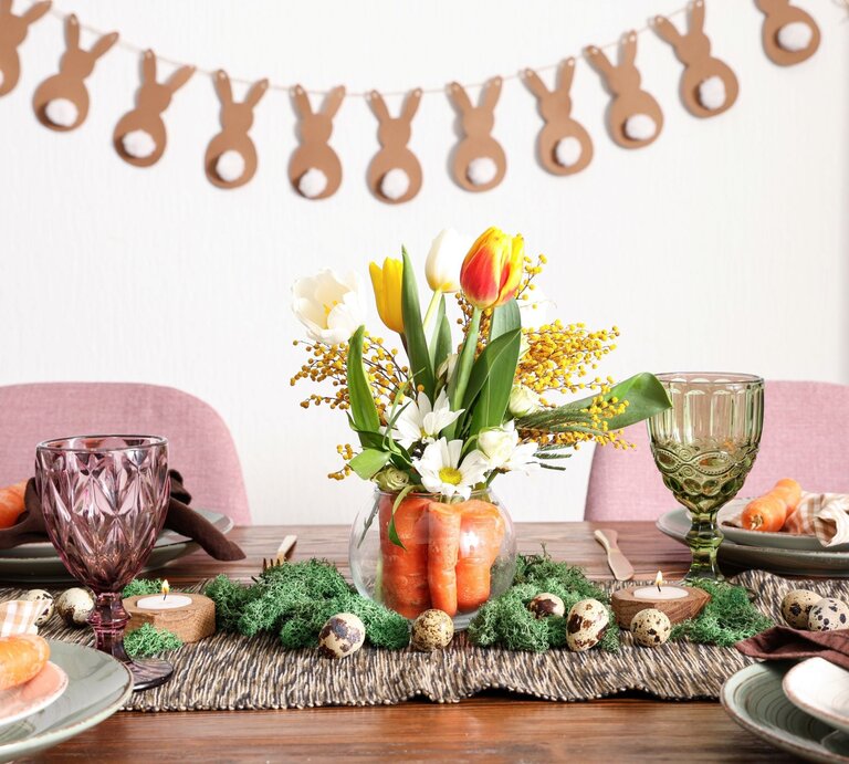 Frühlingshaft gedeckter Tisch mit Tulpenstrauß in Karottenvase, bunten Gläsern, Möhren auf Tellern und Hasengirlande im Hintergrund. | © Shutterstock/Pixel-Shot