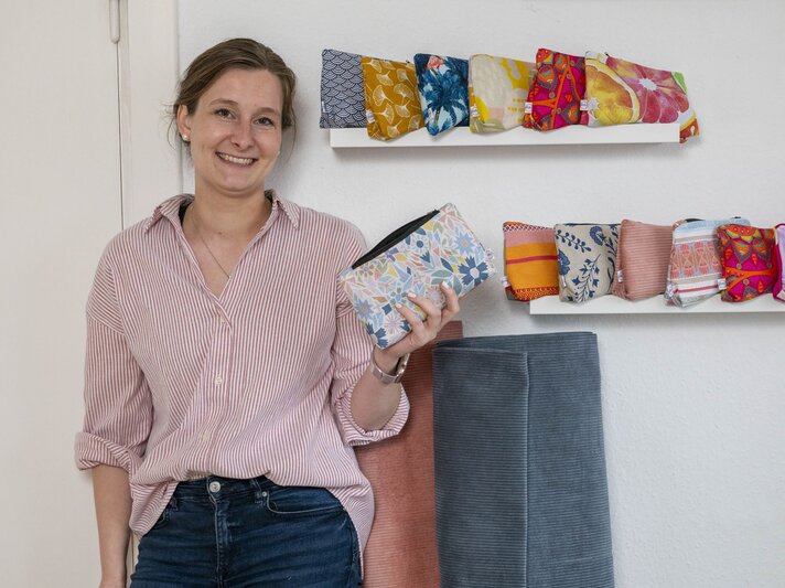 Eine lächelnde Frau mit hochgesteckten Haaren, gestreifter Bluse und Jeans hält eine Stofftasche vor einem Regal mit bunten Täschchen. | © BILD der FRAU