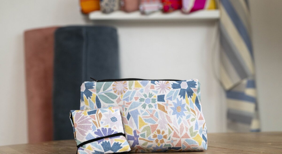 Zwei Kosmetiktaschen mit floralen Mustern in Pastellfarben stehen auf einem Holztisch, im Hintergrund Stoffe und Deko.